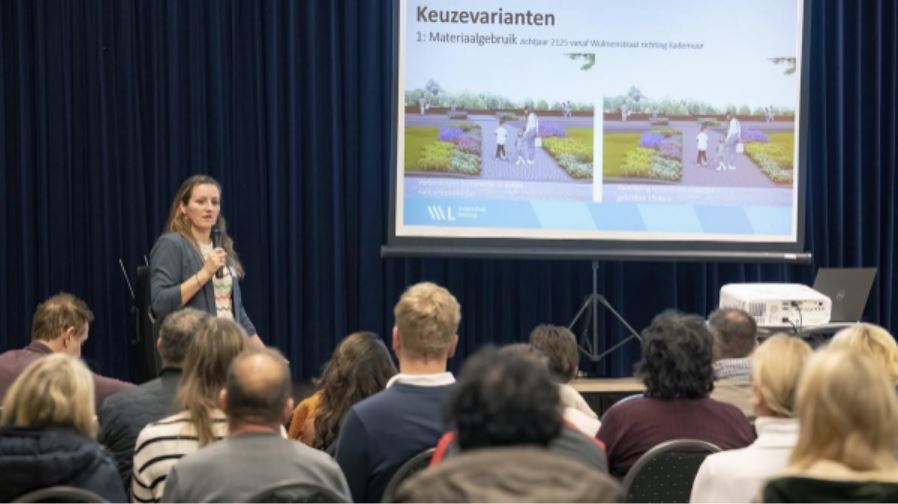 Foto van een vrouw met microfoon in hand die een presentatie geeft aan een publiek